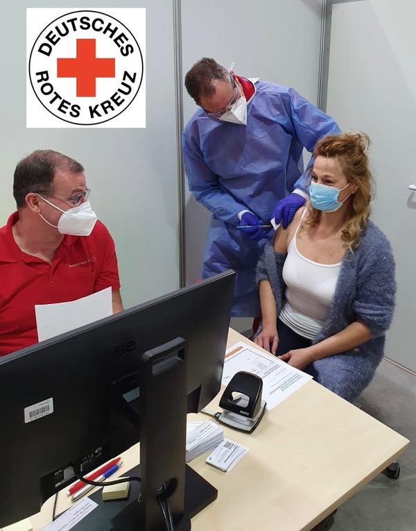 Auf dem Bild sind drei Menschen zu sehen: eine Patientin, die geimpft wird, eine Fachkraft, die die Impfung verabreicht und ein weiterer Mitarbeiter vor einem PC.