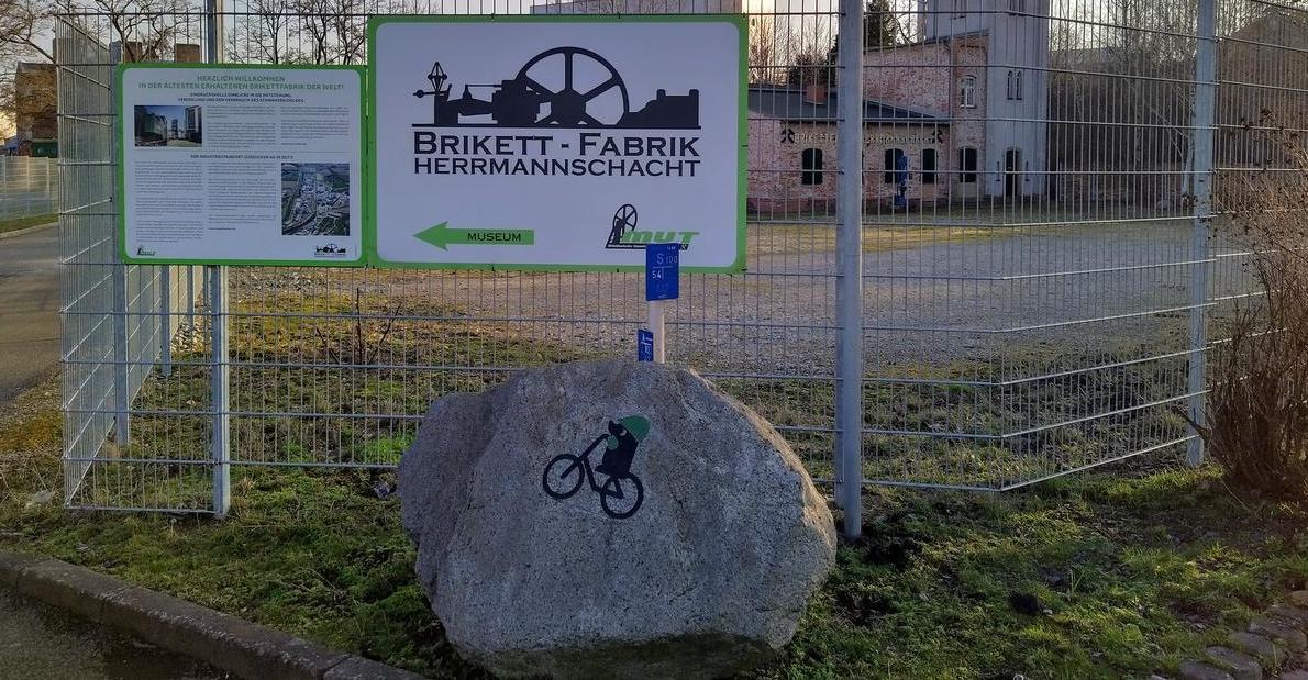 Stein mit der Kennzeichnung des Recoarbo-Kohleradwegs vor einm Schild der Brikettfabrik Herrmannschacht