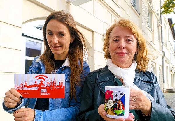 Zwei Mitarbeiterinnen zeigen mit Gutschein und Tassen mögliche Geschenkideen, die man in der Zeitzer Tourist Information kaufen kann,